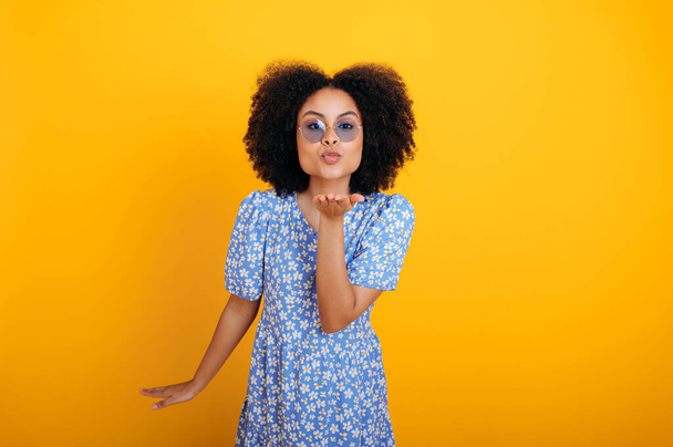 Gelukkig mooi modieus Afrikaans amerikaans krullend behaarde vrouw met een blauwe bril, het dragen van blauwe zomer jurk, poseren, stuurt een luchtkus naar de camera terwijl staan op geïsoleerde oranje achtergrond - Foto, afbeelding