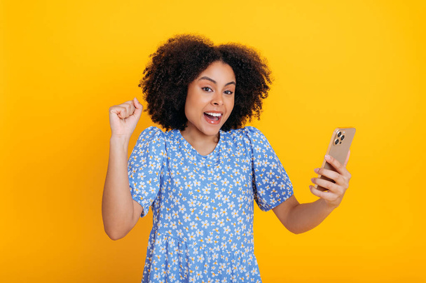 Радісна афроамериканська або бразильська кучерява молода жінка, тримає смартфон в руці, роблячи жест так, радіючи успіху, перемозі, новинам, дивлячись на камеру в подиві, ізольований помаранчевий фон
 - Фото, зображення