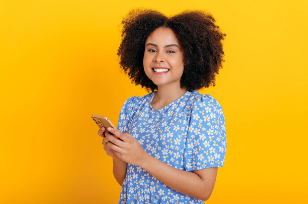 Drahtlose Technologie. Positive afrikanisch-amerikanische oder brasilianische lockige Frau, die Mobiltelefone benutzt, Nachrichten in sozialen Medien sendet, Nachrichten liest, im Internet surft, in die Kamera lächelt, auf orangefarbenem Hintergrund - Foto, Bild