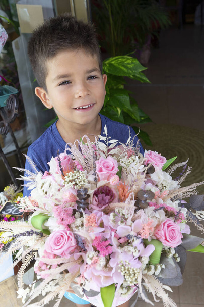Un niño pequeño sonríe encantadoramente con un ramo de flores preservadas en una florería, de cerca. El hombre da flores con rosas. Por un proyecto para el Día de las Madres, y felicitaciones por la fiesta. - Foto, imagen
