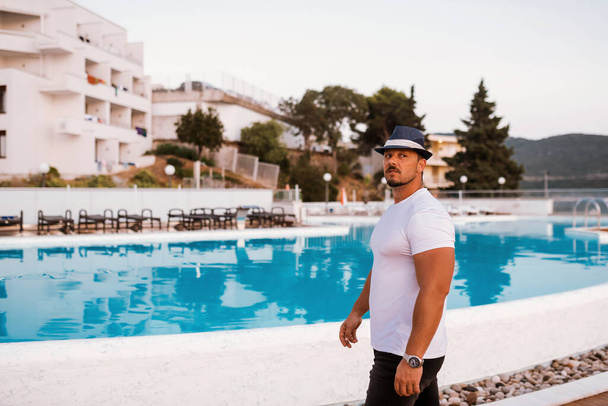 Um homem apto e atraente em férias em um hotel perto de uma praia de areia. Ele está usando um chapéu elegante e desfrutando do sol quente e do mar cristalino. O cenário é sereno e idílico, retratando um sentido - Foto, Imagem