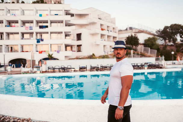 Un hombre en forma y atractivo de vacaciones en un hotel cerca de una playa de arena. Lleva un sombrero elegante y disfruta del cálido sol y el mar cristalino. El paisaje es sereno e idílico, retratando un sentido - Foto, imagen