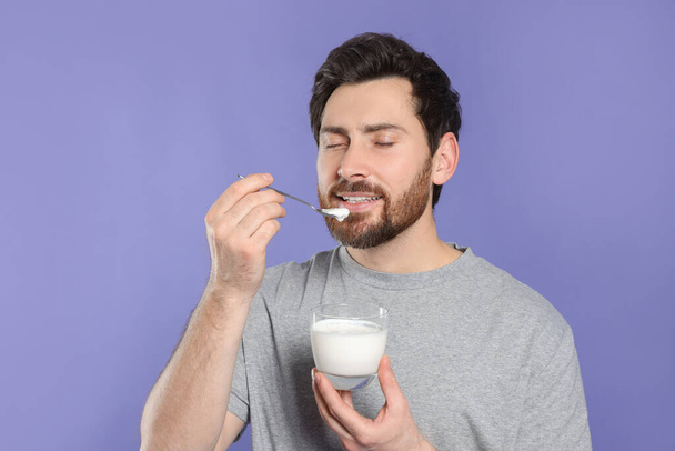 Handsome man eating delicious yogurt on violet background - Фото, изображение
