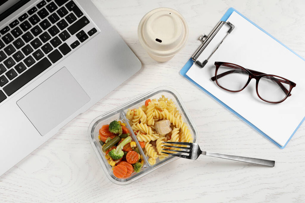 Container of tasty food, fork, laptop, paper cup of coffee and clipboard on white wooden table, flat lay. Business lunch - Фото, изображение