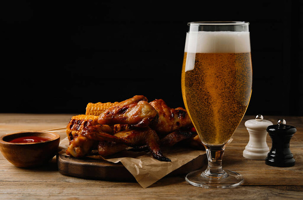 Glass of beer, delicious baked chicken wings, grilled corn and sauce on wooden table against black background - Фото, изображение