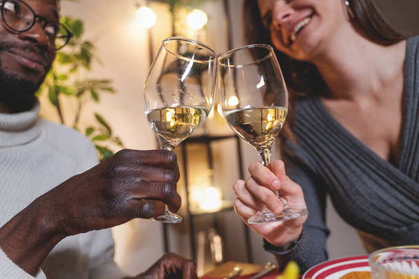 Zbliżenie kieliszków do wina jako międzyrasowej pary - Afrykanin i kobieta kaukaska - tosty z białym winem, ich zamazane twarze śmiejące się w tle w restauracji. - Zdjęcie, obraz