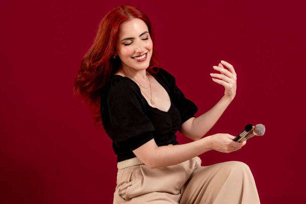 Woman looking at camera and smiling while holding makeup brushes standing against an isolated background. Beauty and cosmetic concept. - Photo, Image