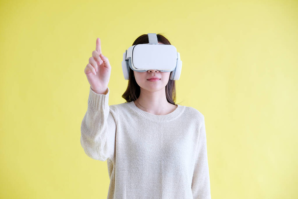 Asian woman wearing virtual reality, VR headset and trying to touch something with hand while stand over isolated yellow background - Fotoğraf, Görsel