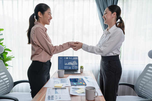 Two Asian businesswomen shaking hands in modern office after successfully analyzing piles of dashboard data papers, as importance of teamwork and integrity in the workplace concept. Enthusiastic - Photo, Image
