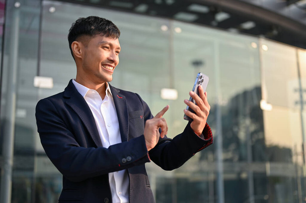 Happy and smiling millennial Asian businessman in a formal business suit using his smartphone while standing outside of the company building. - Fotó, kép
