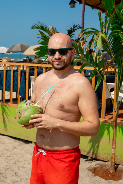 Bald man in sunglasses tries coconut milk from coconut, stands against background of beach. Tourism and travel. Enjoying taste. New sensations. Happiness. Tropical paradise. - Photo, Image