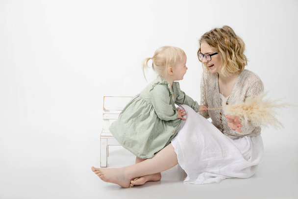 Happy young mother in white dress is sitting on floor, laughing and playing with her little daughter on white background in studio. Love and care. Happy childhood. Child development. Relationship. - Photo, Image