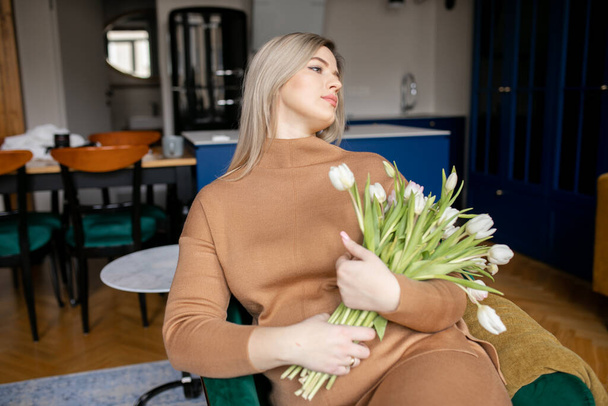 Pregnant young blonde with long hair in beige suit comfortably settled on sofa with bouquet of tulips at home. Modern business ladies. Self-sufficient women. Healthy pregnancy. Lifestyles. - Photo, Image