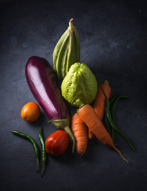 various organic vegetables on a dark textured surface, top view of eggplant, carrots, tomatoes, ridged gourd, chayote and green chilies - Foto, immagini
