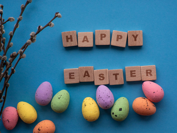 Happy Easter concept. Easter decorations on blue background with copy space. Easter greating card. Flat lay. Colorful eggs and Happy Easter inscription on wooden cubes. Celebration of spring holidays. - Foto, Imagen