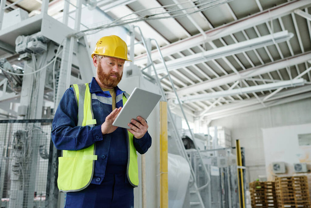 Mladý muž inženýr s tabletem se zaměřením na čtení online informací, zatímco stojí v prostorné dílně továrny - Fotografie, Obrázek