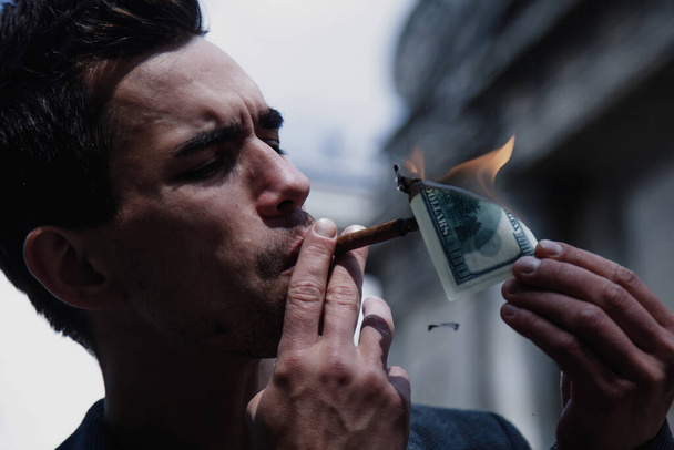 Junger brutaler Geschäftsmann zündet Zigarre mit 100-Dollar-Schein an. Horizontales Bild.  - Foto, Bild