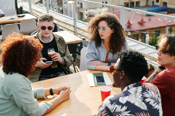 Gruppe junger interkultureller Freunde in stylischer Freizeitkleidung, die sich im Café im Freien oder auf dem Dach ausruhen, während sie am Tisch sitzen und plaudern - Foto, Bild