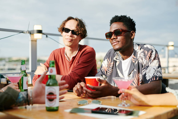 Deux mecs interculturels en lunettes de soleil et vêtements décontractés prenant des boissons dans un café en plein air assis à table devant leurs amis pendant le chat - Photo, image