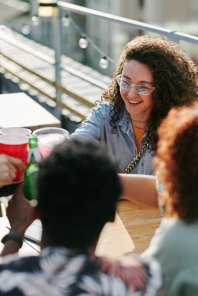 Молода весела жінка в окулярах і повсякденному одязі тости зі своїми друзями зібралися за столом у відкритому кафе на літніх вихідних
 - Фото, зображення