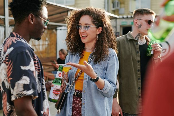Ευτυχισμένο κορίτσι κρατώντας ένα ποτήρι μαρτίνι με κοκτέιλ και μιλώντας με μαύρο τύπο με ένα μπουκάλι μπύρα απολαμβάνοντας το κόμμα ή τη συγκέντρωση των φίλων - Φωτογραφία, εικόνα