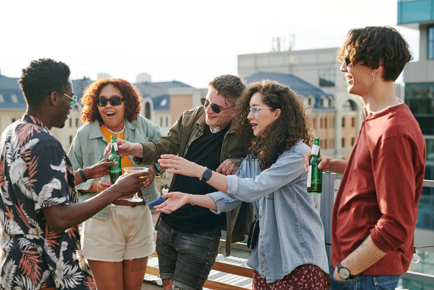 Young black man passing bottle of beer and cocktail to guy and girl during rooftop party or gathering in outdoor cafe on weekend - Photo, Image