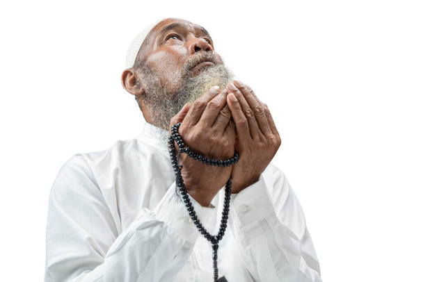 Homme musulman avec une barbe portant une casquette blanche priant avec des perles de prière sur les mains tout en étant assis isolé sur fond blanc - Photo, image