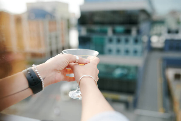 Hands of young woman holding martini glass with cocktail while having rest on terrace or rooftop cafe against urban environment - Foto, immagini