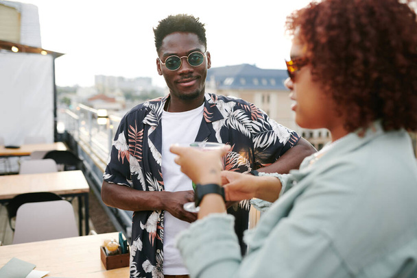 Güneş gözlüklü Afro-Amerikalı bir adam kız arkadaşının önünde duruyor ve onunla sohbet ederken ikisi de çatıdaki kafede içki içiyor. - Fotoğraf, Görsel