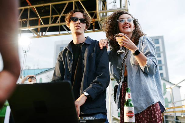 Glückliche junge Frau mit Cocktail, die neben ihrem Freund steht oder als DJ Musik am Plattenteller für Freunde auf einer Dachparty macht - Foto, Bild