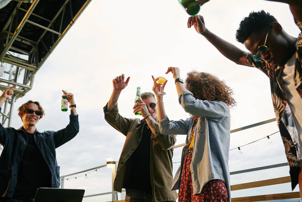 Grupa młodych ekstatycznych przyjaciół z butelkami piwa tańczących na imprezie na dachu w kawiarni na świeżym powietrzu, stojąc z podniesionymi ramionami - Zdjęcie, obraz