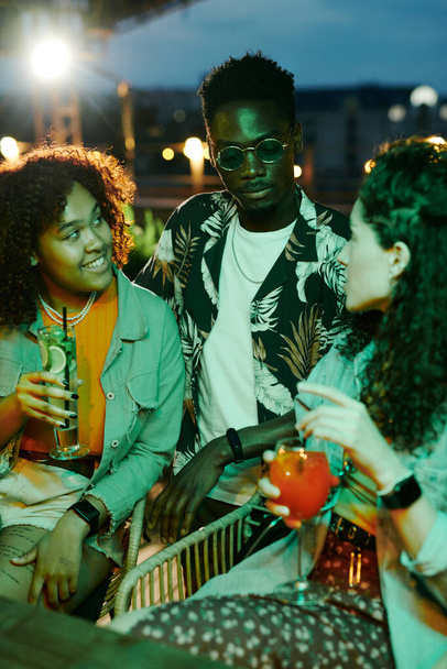 Zwei interkulturelle Mädchen mit Cocktails schauen einen Afroamerikaner in stylischer Freizeitbekleidung und Sonnenbrille an, der zwischen ihnen sitzt. - Foto, Bild