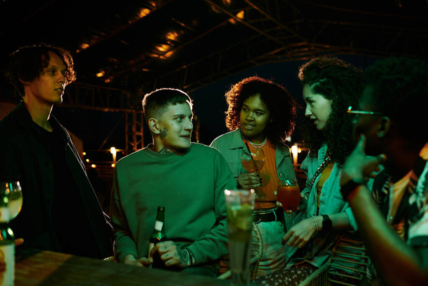 Youthful intercultural girls with cocktails and guys listening to young man with bottle of beer while enjoying party in night club - Foto, afbeelding