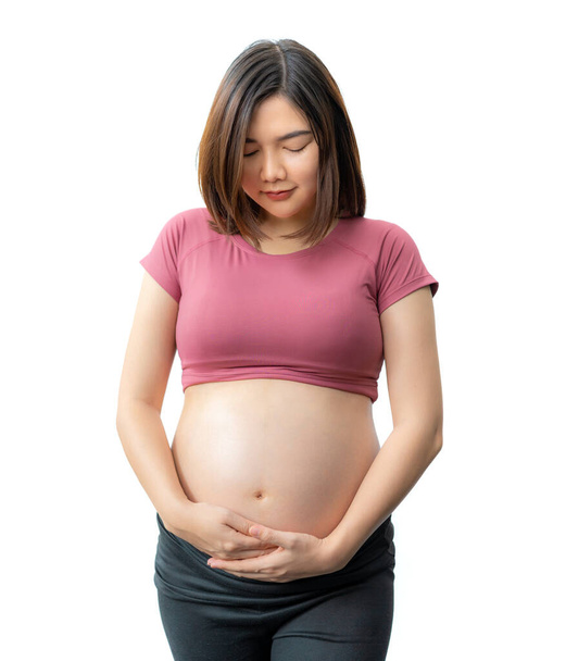 Happy smiling Asian pregnant woman in fitness clothes standing holding her belly against white background, room for copy space - Foto, Bild