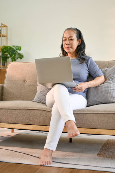 Retrato de una mujer jubilada feliz de edad asiática en ropa casual usando su computadora portátil en el sofá de su sala de estar. - Foto, Imagen