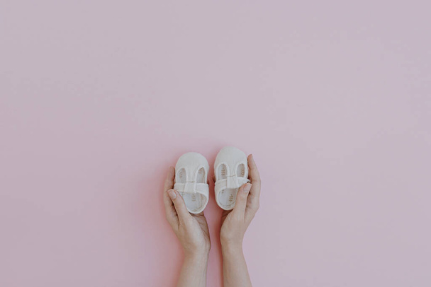 Person's hands hold cute little baby sandals shoes on pastel pink background. Minimalist baby fashion apparel - Photo, Image