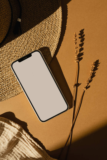 Smartphone met blanco kopieerruimte scherm, stijlvolle strohoed, droog gras op warme bruine achtergrond. Esthetisch zonlicht schaduw silhouet. Online winkelen concept. Luxe Boheemse minimalistische achtergrond - Foto, afbeelding