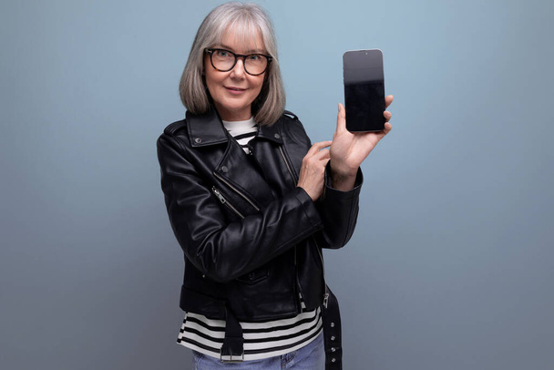 dromerige moderne jaren '60 moderne oude dame met grijs haar houdt een apparaat verticaal met een mockup op een heldere achtergrond met kopieerruimte. - Foto, afbeelding