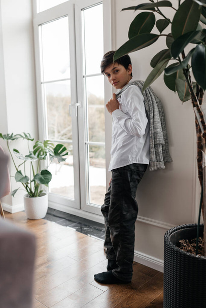 portrait of a pretty teenager in white shirt standing near the window indoors, holding sweater on his shoulder and smiling at camera.  - Fotografie, Obrázek