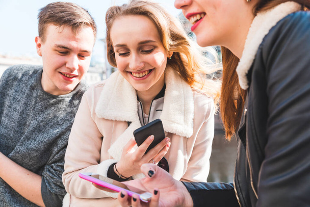 Happy group of friends using smartphone and laughing - Three young people, a man and two women, relaxing and having fun, smiling and talking together - Friendship and lifestyle concepts - Fotó, kép