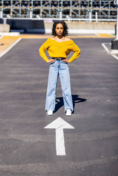 Woman portrait standing in front of an arrow pointing at her - Beautiful multiracial young woman wearing yellow top and curly hair - determination and lifestyle concepts - Φωτογραφία, εικόνα