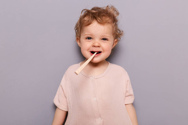 Image of sweet baby girl with blond hair. brushes her teeth with smile, dental health taking care of mouth hygiene, having fun, standing isolated over gray background. - Photo, image