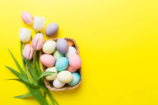 Happy Easter composition. Easter eggs in basket on colored table with yellow Tulips. Natural dyed colorful eggs background top view with copy space. - 写真・画像