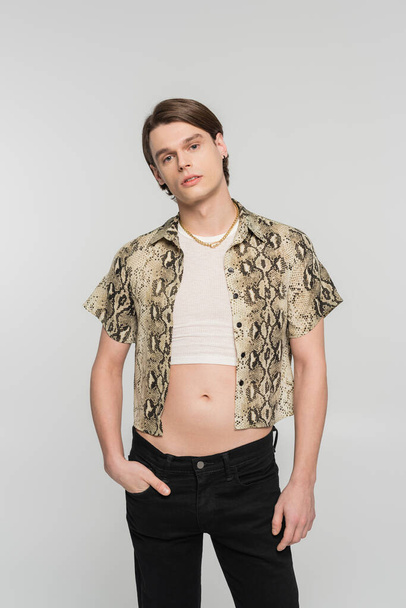 stylish bigender person in snakeskin print blouse posing with hand in pocket of black pants isolated on grey - Фото, изображение