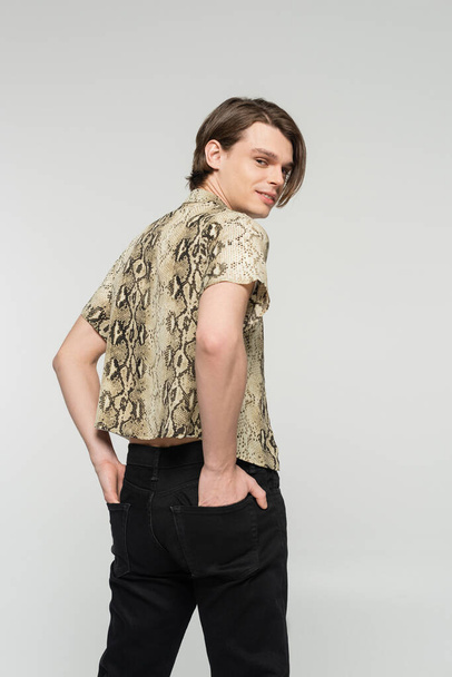 trendy pangender person in animal print blouse holding hands in back pockets and smiling at camera isolated on grey - Photo, Image