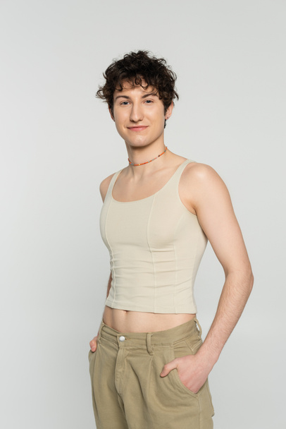 brunette bigender person in tank top standing with hands in pockets of beige pants and smiling at camera isolated on grey - Fotoğraf, Görsel