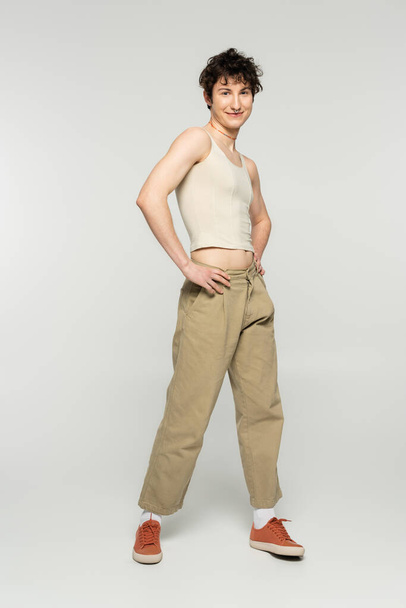 full length of happy bigender model in beige pants posing with hands on hips on grey background - 写真・画像