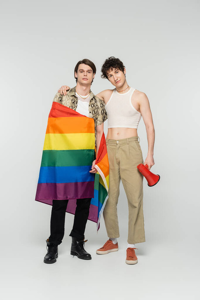 full length of young nonbinary couple with lgbt flag and megaphone looking at camera on grey background - Photo, Image