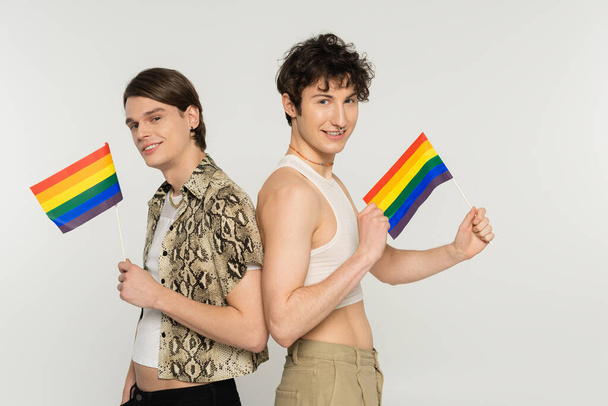 trendy and carefree pansexual couple standing with small lgbt flags and smiling at camera isolated on grey - Foto, Bild