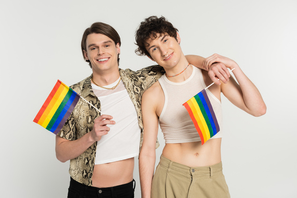 young and carefree bigender couple with rainbow flags embracing and smiling at camera isolated on grey - Fotografie, Obrázek